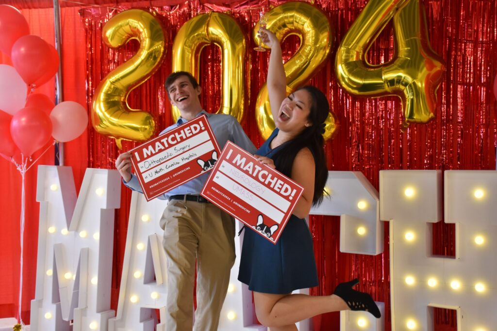 Man and woman in front of bright red background white match letters celebrating with their I matched signs