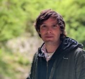 head and shoulders shot of andrew ellison outside in a rural environment with green plants surrounding him, wearing a dark gray jacket.