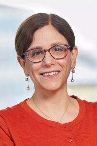 Head and shoulders of Dr. Bair-Merritt wearing red top, pearl earrings, brown eyeglasses, dark brown hair, smiling