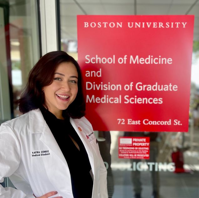 Kayra Cengiz wearing whitecoat in front of red sign for BU GMS and Chobanian & Avedisian SOM