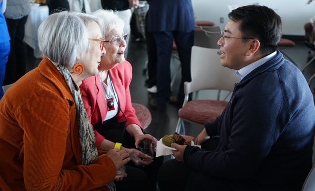 Two women with silver/gray hair seated and talking young man with dark short hair and eyeglasses