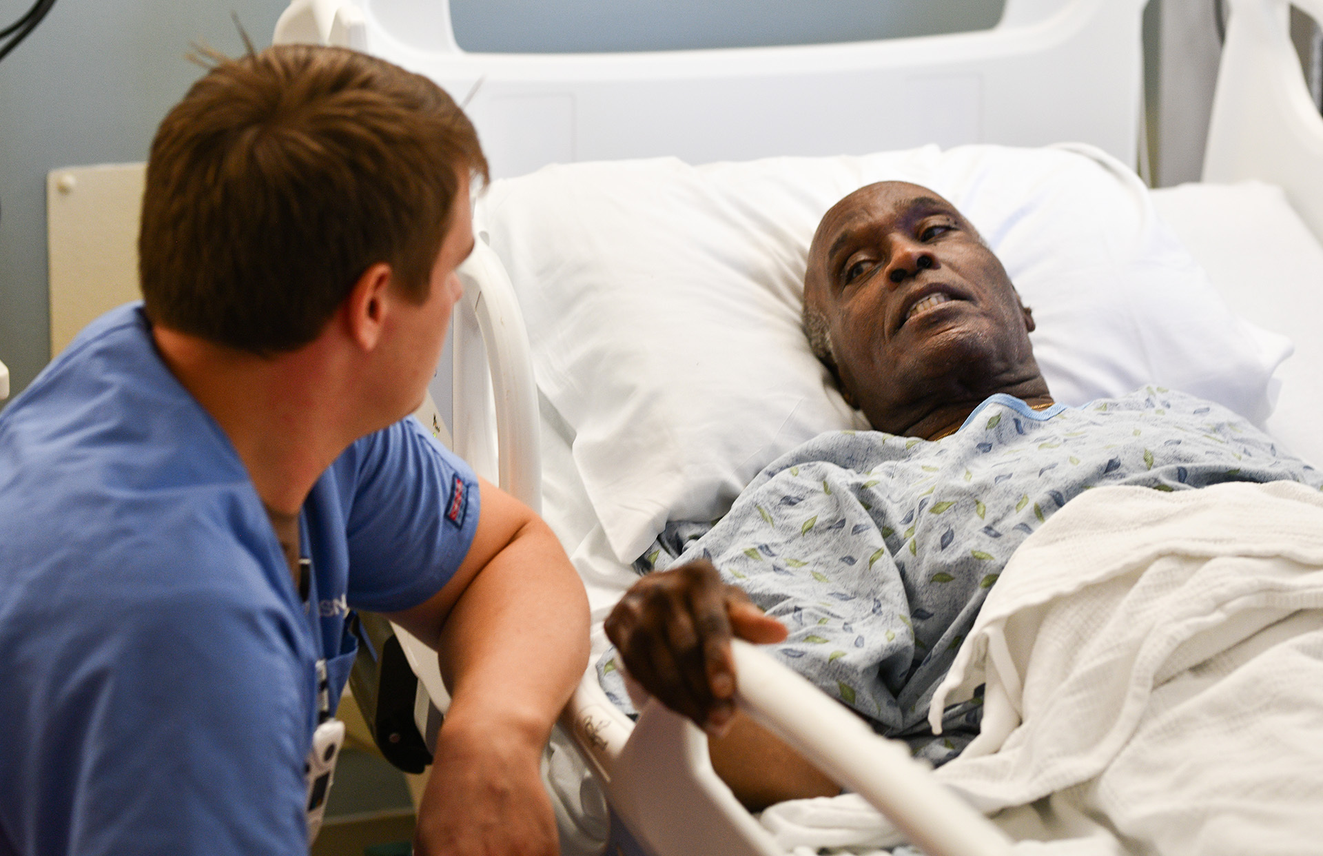 Medical student in blue scrubs seated next to patient lying on bed