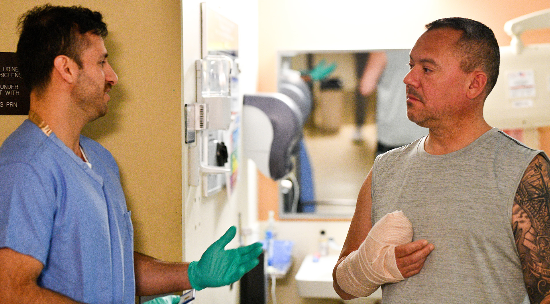 Medical student in blue scrubs and green gloves talking with man patient with heavily bandaged hand standing in hospital hallway