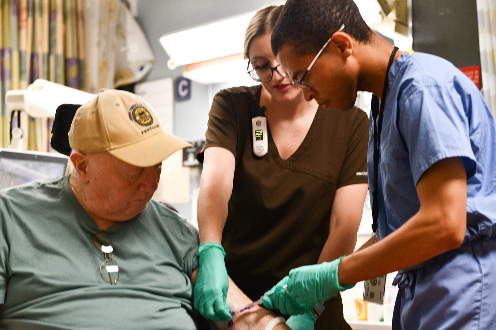 Man medical student in blue scrubs and green gloves flushing man patient's IV line with nurse in brown scrubs and green gloves directing and medical student