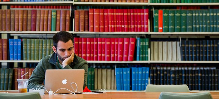 A student working on a laptop in a library, surrounded by bookshelves.