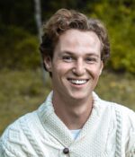 Man with light brown wavy hair wearing white heavy knit sweater smiling outside in area with green field in background