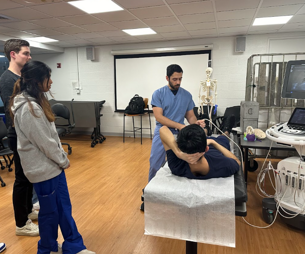 A medical instructor performs an ultrasound while two students observe in a clinical classroom setting.