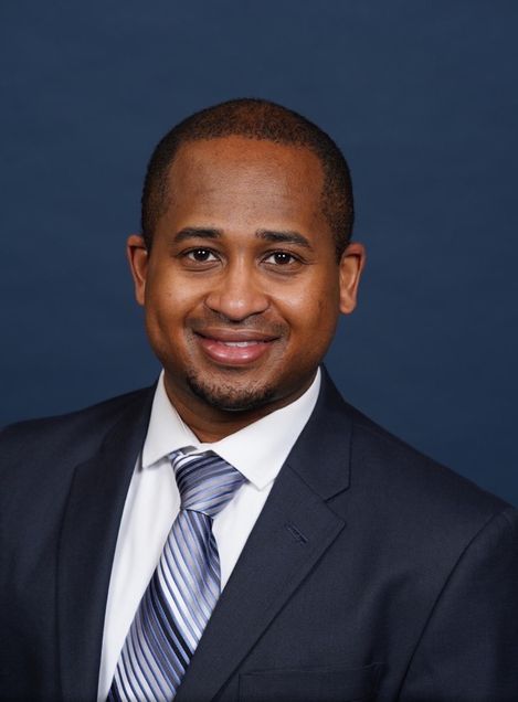 Portrait of Dennis Jones wearing dark blue suit jacket, white sirt, blue and gray necktie, smiling, short dark hair
