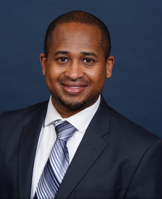 Portrait of Dennis Jones wearing dark blue suit jacket, white sirt, blue and gray necktie, smiling, short dark hair