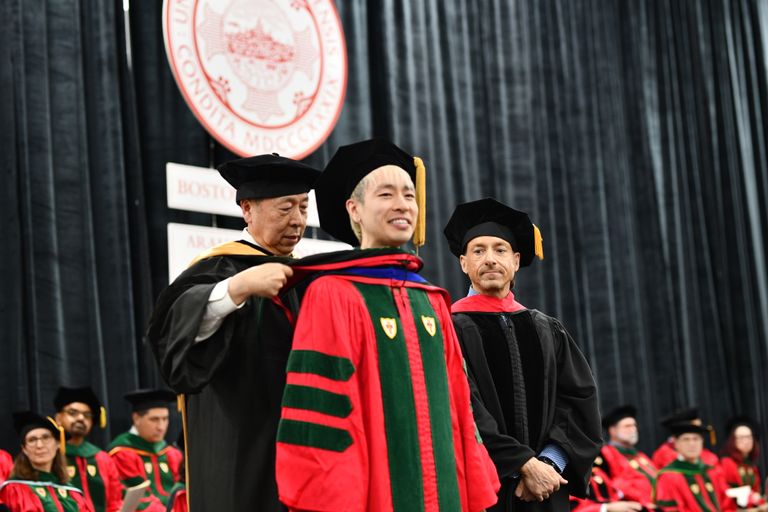 Graduate being hooded by a faculty member at graduation ceremony with BU emblem in the background..