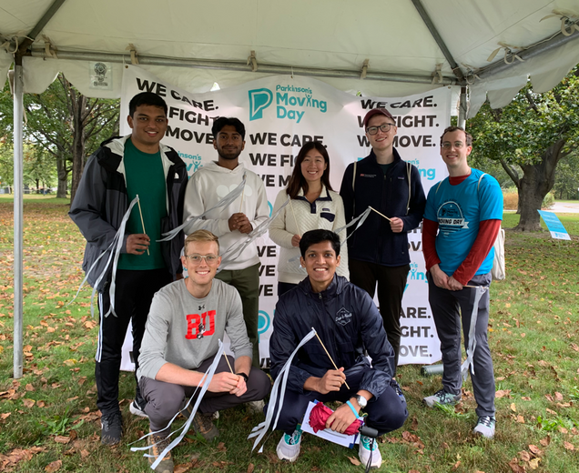 A group of seven people posing in front of a "Parkinson's Moving Day" banner under a tent, holding white ribbons.
