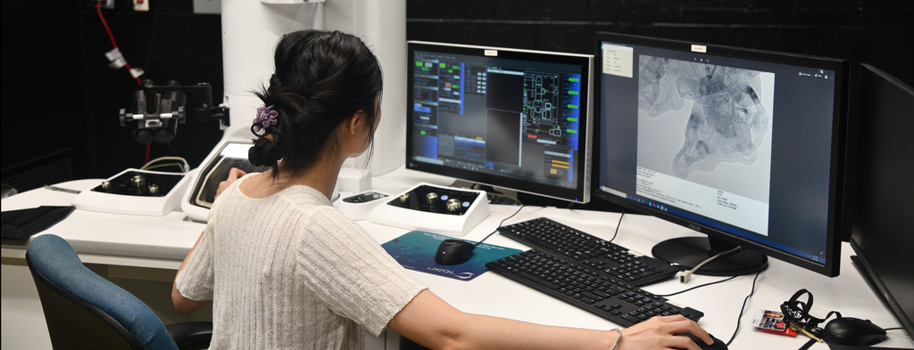 A person analyzing medical imaging on multiple computer monitors in a lab setting, with advanced equipment visible on the desk.