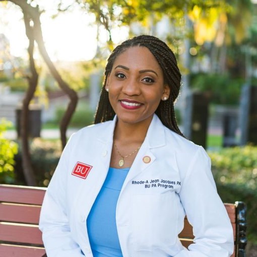 Rhode-Armelle Jean Jacques wearing a lab coat and a blue blouse, standing outside with trees and sunlight in the background
