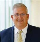 head and shoulders image of Mark Conrad, short gray heair eyeglasses, broad smile, wearing white shirt, yellow tie, blue suitjacket