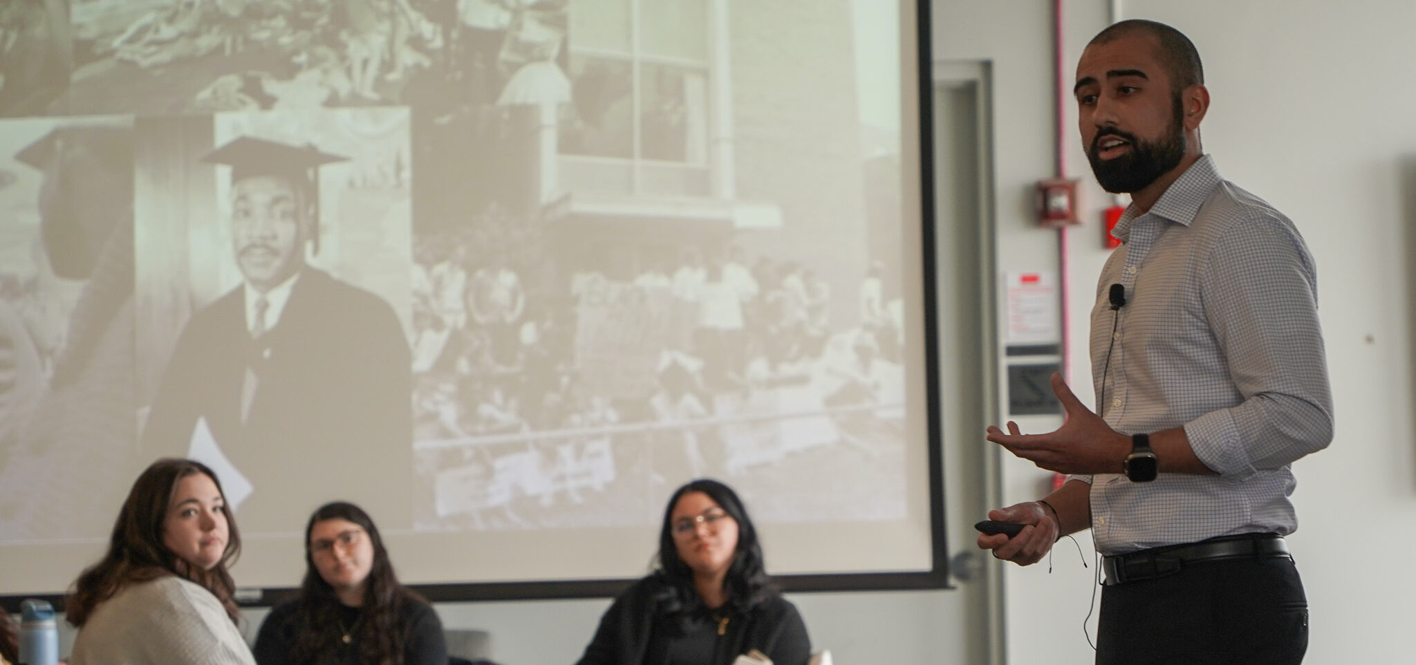 neil Bedi standing and lecturing infront of seated faculty, staff, students. MLK images in background.