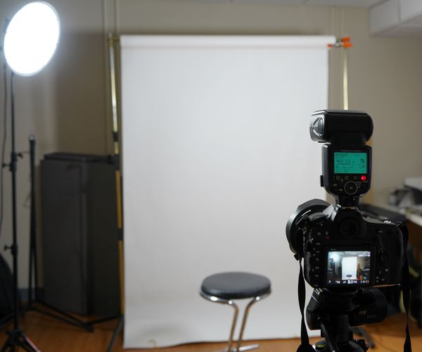 A professional video recording studio setup with a camera mounted on a tripod facing a stool, positioned in front of a white backdrop and a large circular light source is placed to the left of the camera