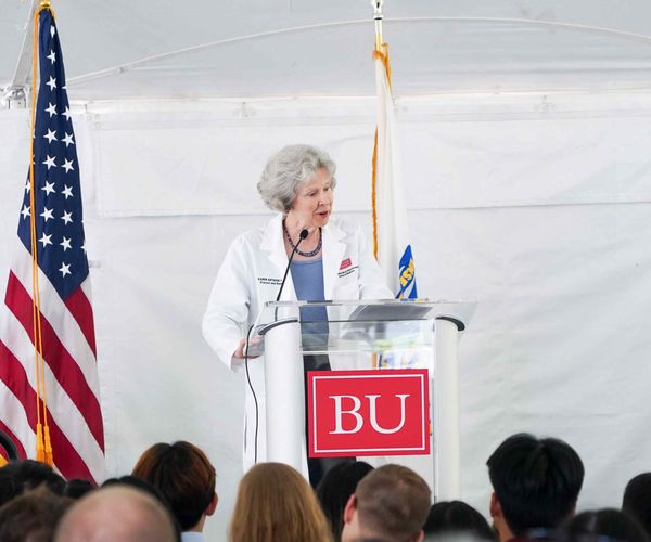 A person in a white coat standing at a podium with a Boston University logo, with American and Boston flags in the background, speaking to an audience