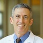 head and shoulders shot of Eric Schwartz, gray hair, smiling broadly, wearing blue shirt, dark necktie and whitecoat
