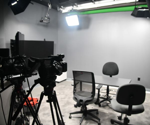 A professional video recording studio setup with multiple cameras on tripods, office chairs around a table, a partially visible green screen, and overhead lights, including a large softbox