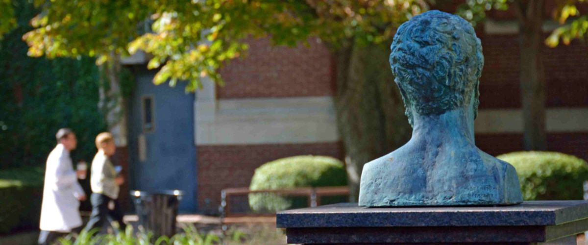 A bronze or copper statue of a person's head and shoulders on a pedestal, with people walking and a brick building in the background