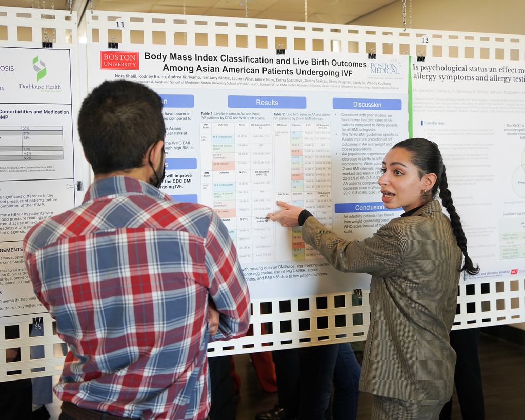 Medical student Nora Khalil standing in front of large poster pointing with left hand looking at man wearing a plaid shirt appears to be listening.
