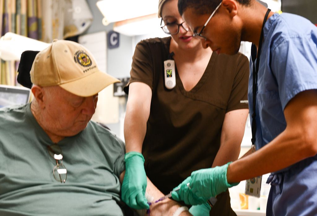 Second-year med student Douglass Bryant, working under supervision of vascular access nurse Kassidy Dias, RN, flushes an IV line of Army veteran Robert McAllister.