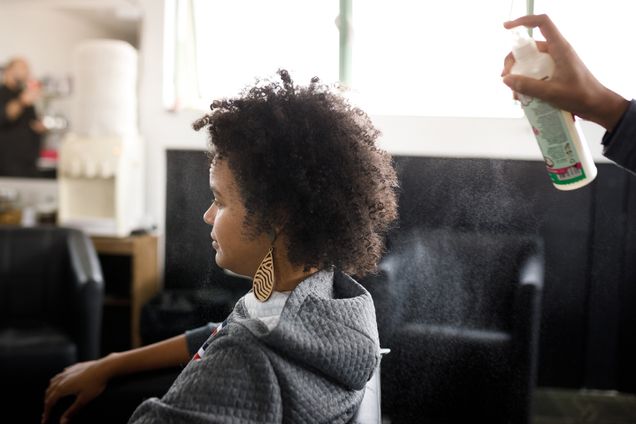 A black woman with an afro sits in a daylight-lit living room space with a person slightly off-camera entering the frame with a hair spray from behind. The photo is shot from a landscape angle to the side of both.
