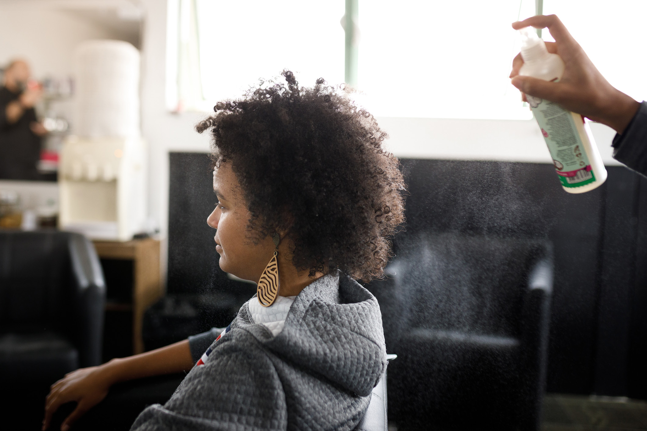 A black woman with an afro sits in a daylight-lit living room space with a person slightly off-camera entering the frame with a hair spray from behind. The photo is shot from a landscape angle to the side of both.