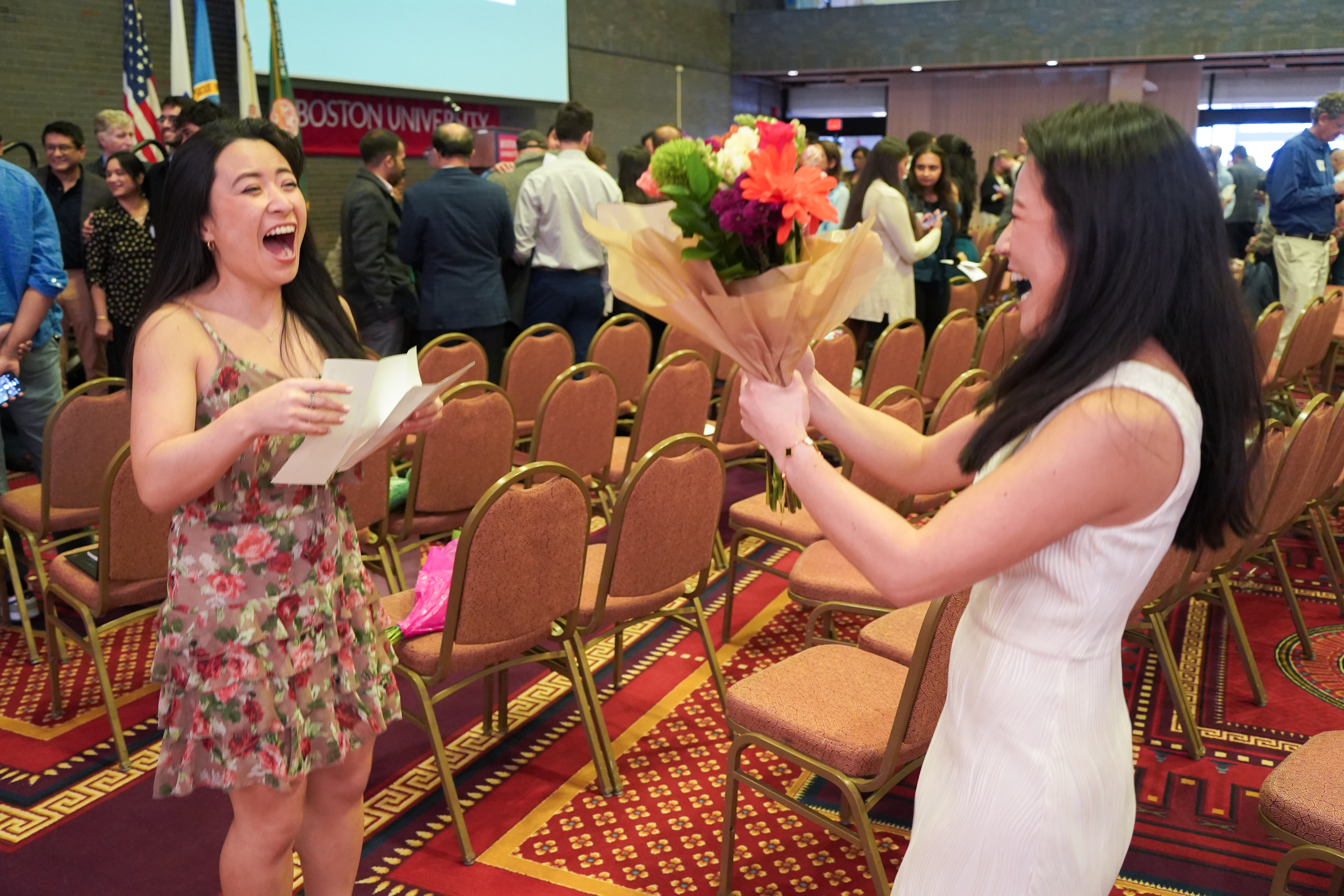 Two woman facing one another smiling broadly one holding flowers, one holding a white paper with Match Day results