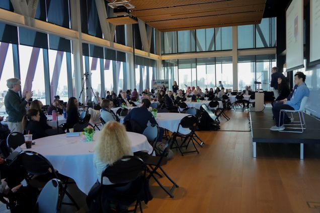 Large room with lots of windows, many large tables with white tablecloths, people sitting and listening to speaker