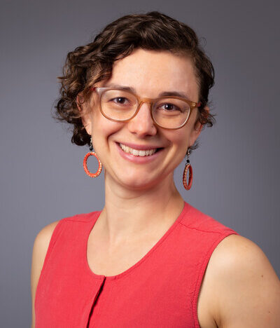 head and shoulders of woman with short brown curly hair, eye glasses, smiling broadly, wearing orange top