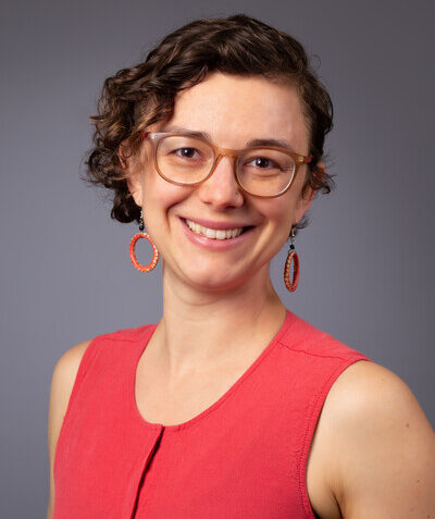 Head and shoulders of Maura Murray Horwitz, short brown curly hair, eyeglasses, smiling broadly, wearing orange top
