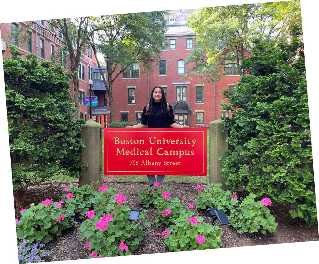 Vejar standing in front of a red sign that says Boston University Medical Campus in a flowering garden