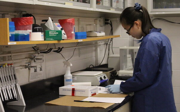 student wearing blue labcoat and gloves at lab bench
