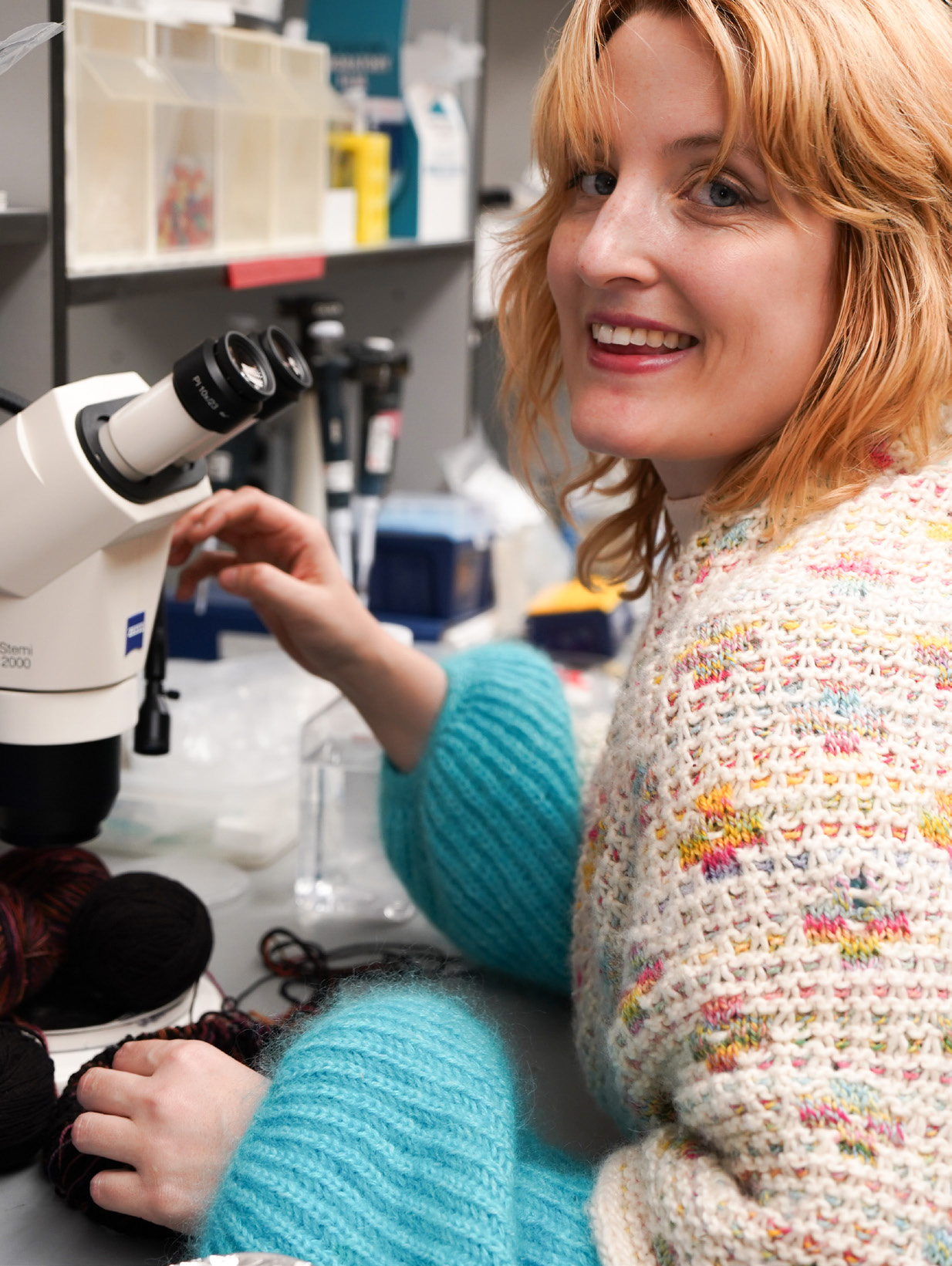 A blond student sits at a microscope, wearing a knitted sweater