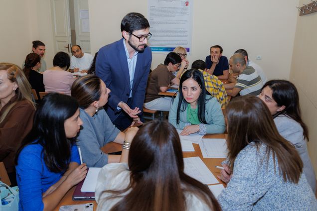 Man in blue jacket stading around tables with papers on them and students seated.