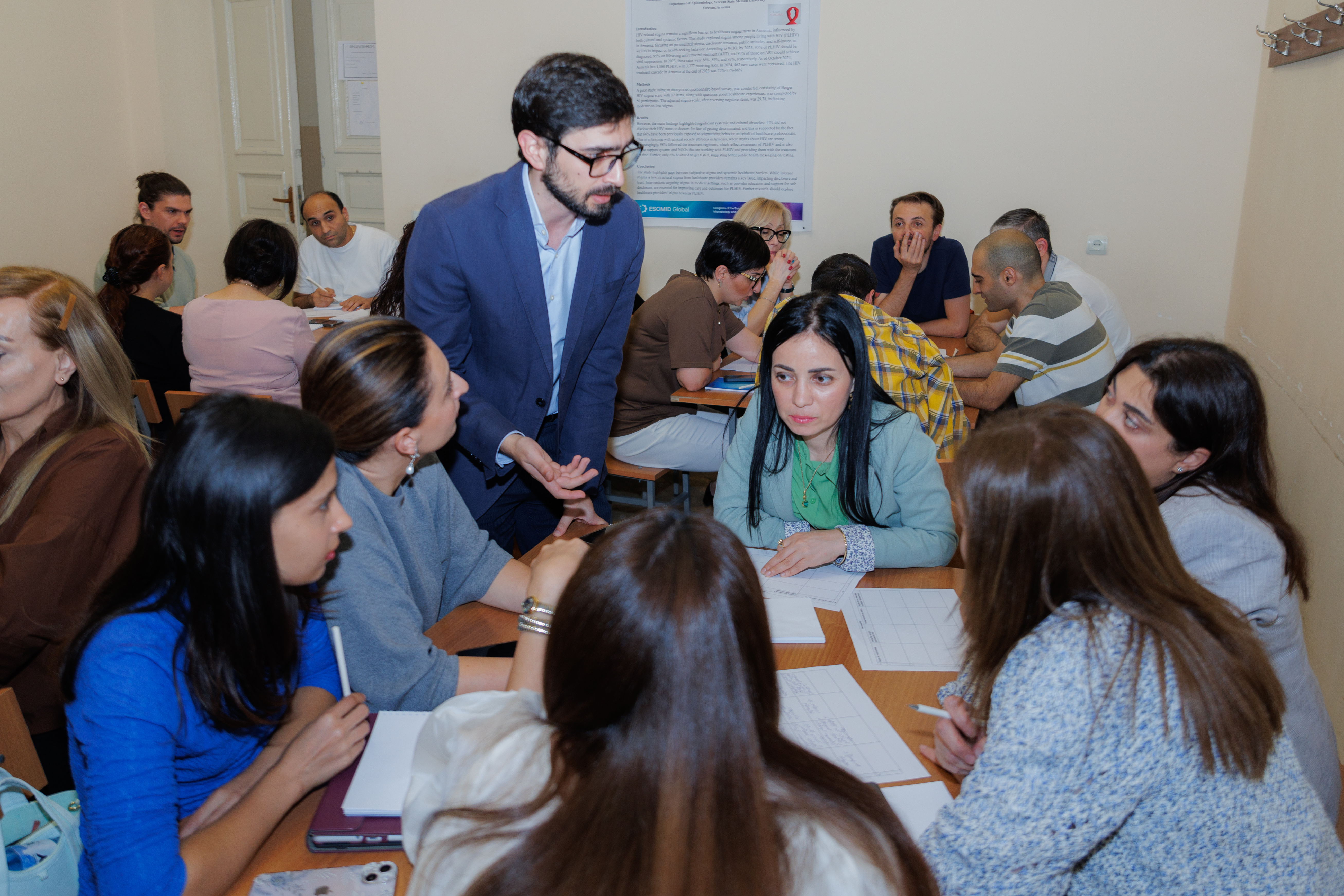 Man in blue jacket stading around tables with papers on them and students seated.