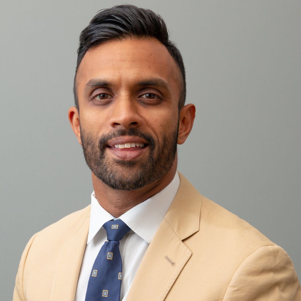 Head and shoulders of man with dark facial hair wearing tan suit jacket and white shirt