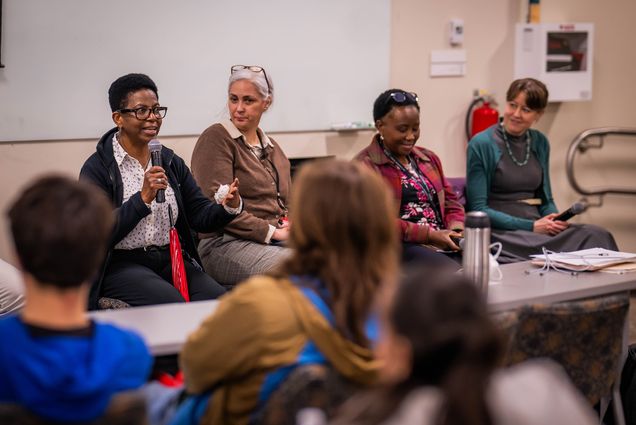 Four people seated at a table speaking to a group of high school students