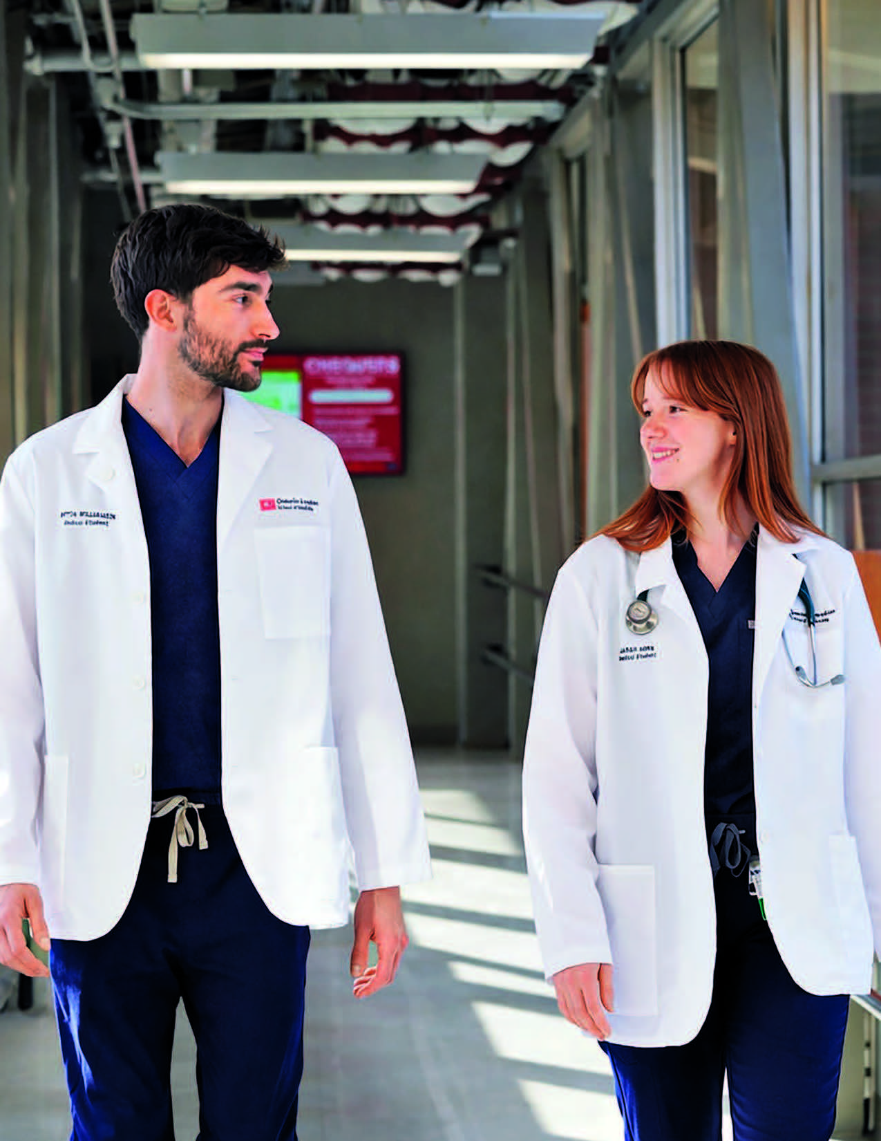 Two students wearing white coats walk down a hallway and look at each other