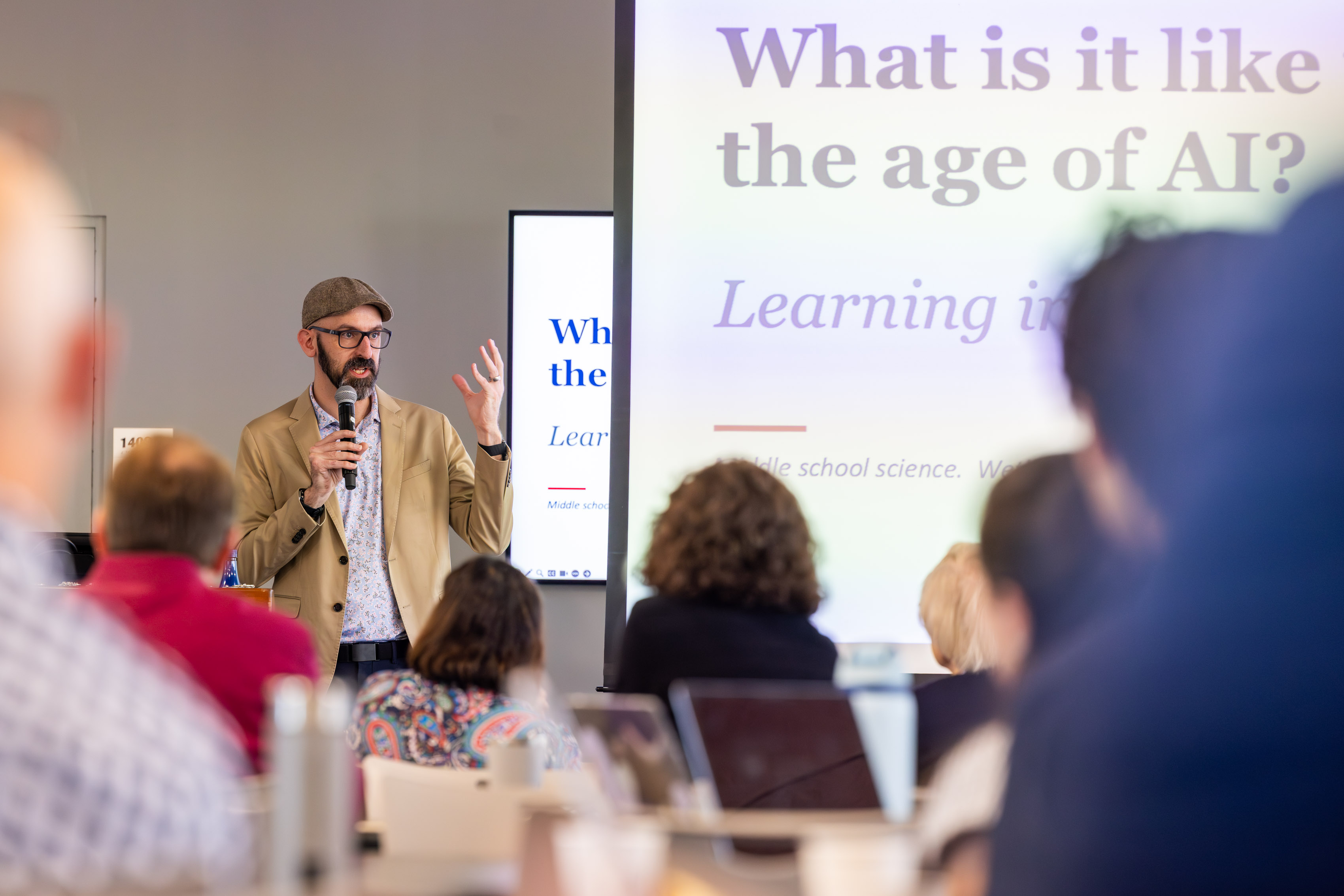 Man with hat and beard speaking to crowd with large powerpoint screens about AI