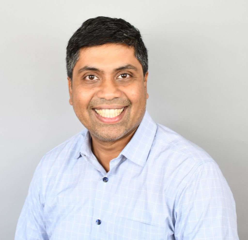 Head and shoulders of man with short dark hair wearing light blue shirt, smiling broadly