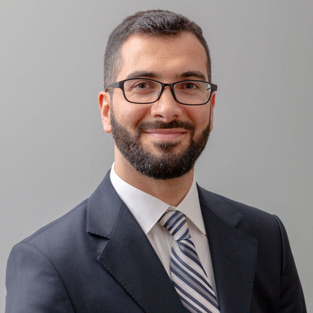 Man with dark hair and facial hair, eyeglasses, dark suit, light shirt, striped tie