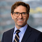 Head and shoulders of man with brown hair, eyeglasses wearing dark suit blue shirt striped blue red white tie