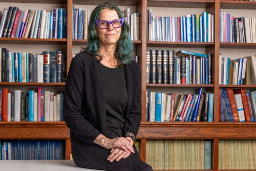 Woman in black with long greenish/blue hair wearing eyeglasses in front of bookcases