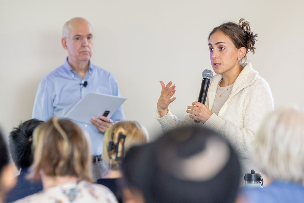 Woman holding microphone standing next to man holding papers, people seated listening