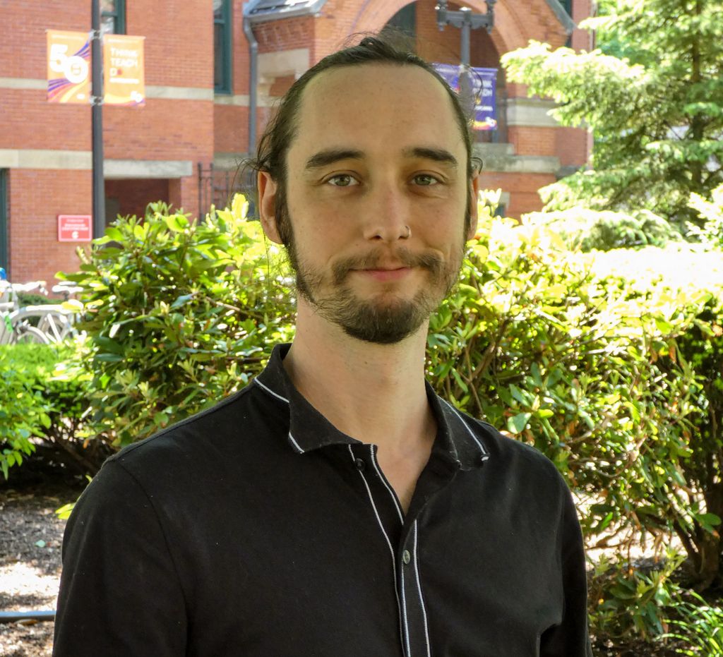 Head and shoulders of man with dark hair, facial hair standing outside in front of Talbot building