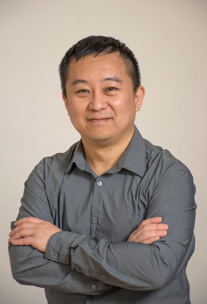 Man with short dark hair, wearing gray shirt, arms folded, gently smiling
