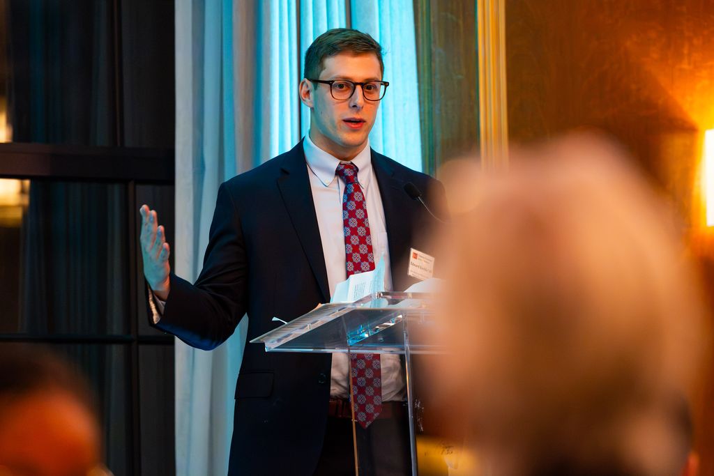Man wearing suit and eyeglasses speaking at podium