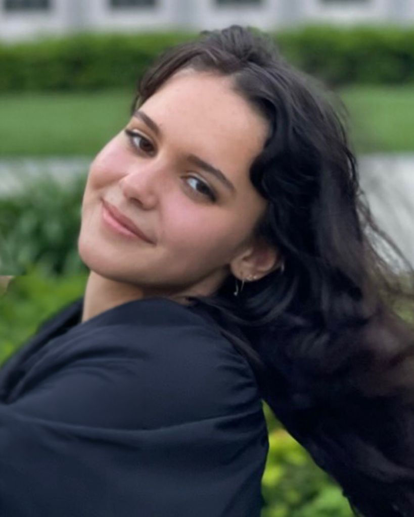 Woman with long dark hair wearing dark top, leaning back outside with grass behind her